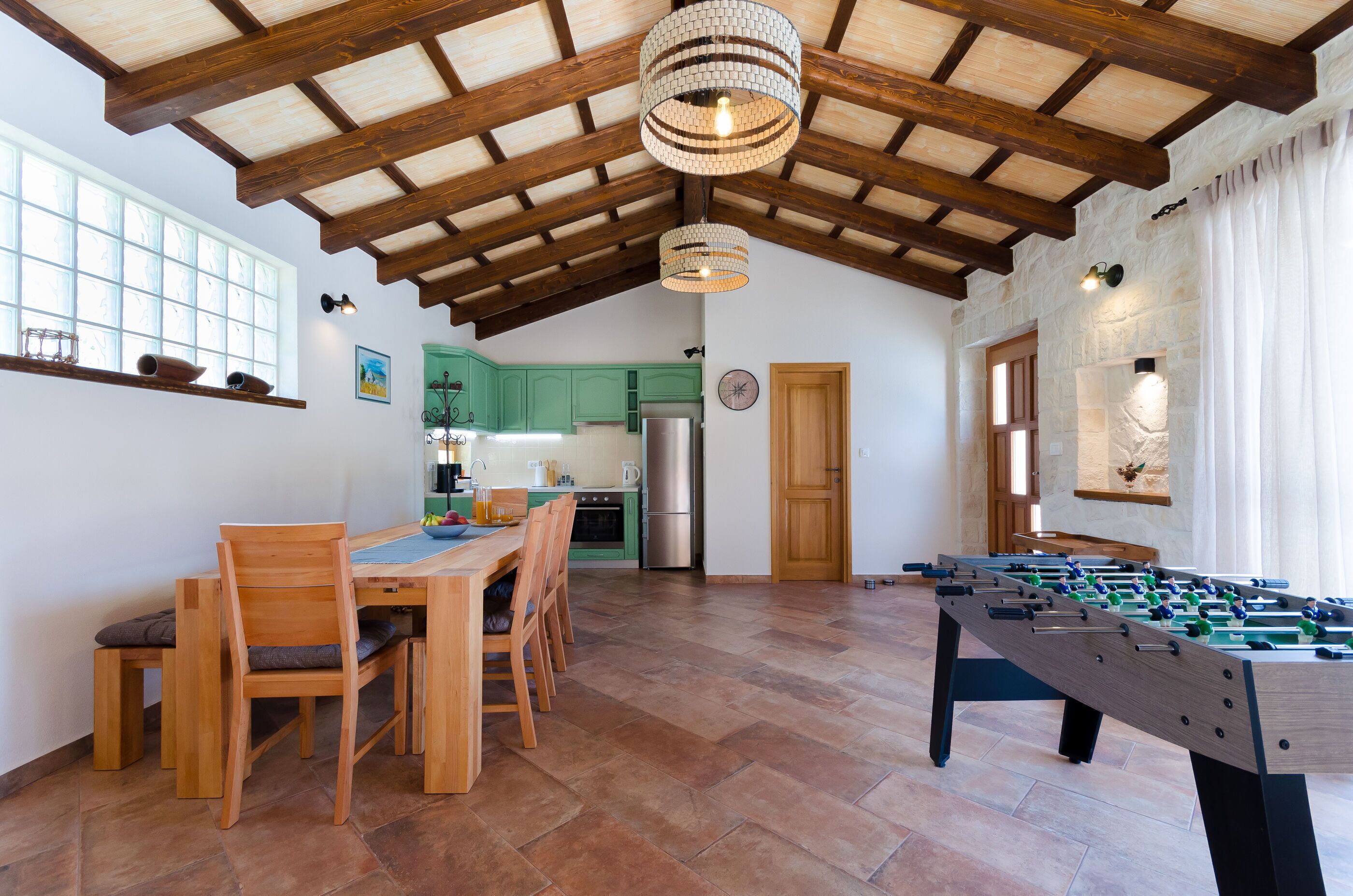 A spacious room with a wooden table and chairs, a foosball table, and a kitchen area with green cabinets.