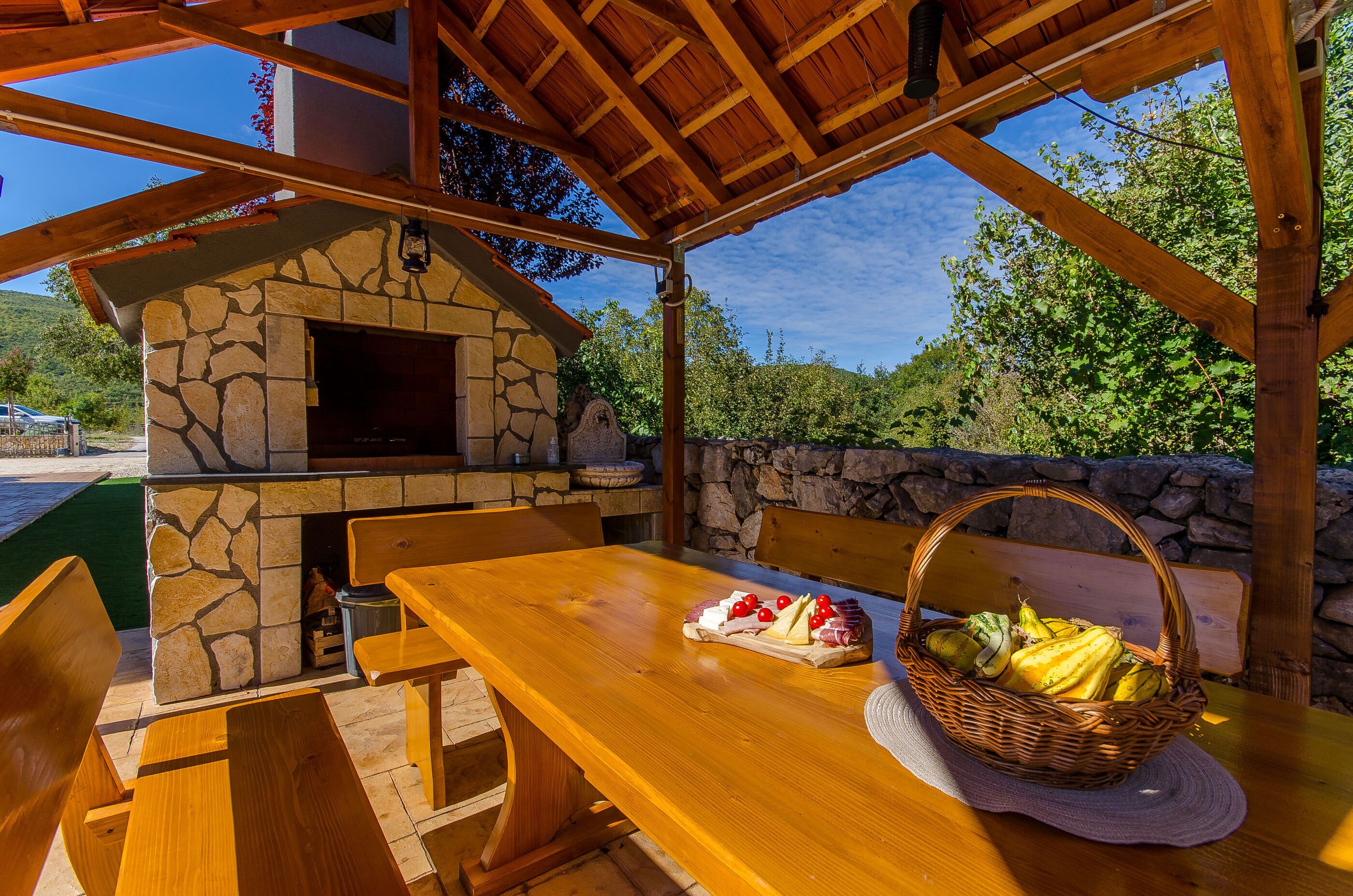 Uma área de refeições ao ar livre com madeira, uma lareira de pedra, uma mesa com cesta de frutas e um painel de parede de pedra.