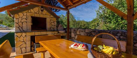 Uma área de refeições ao ar livre com madeira, uma lareira de pedra, uma mesa com cesta de frutas e um painel de parede de pedra.