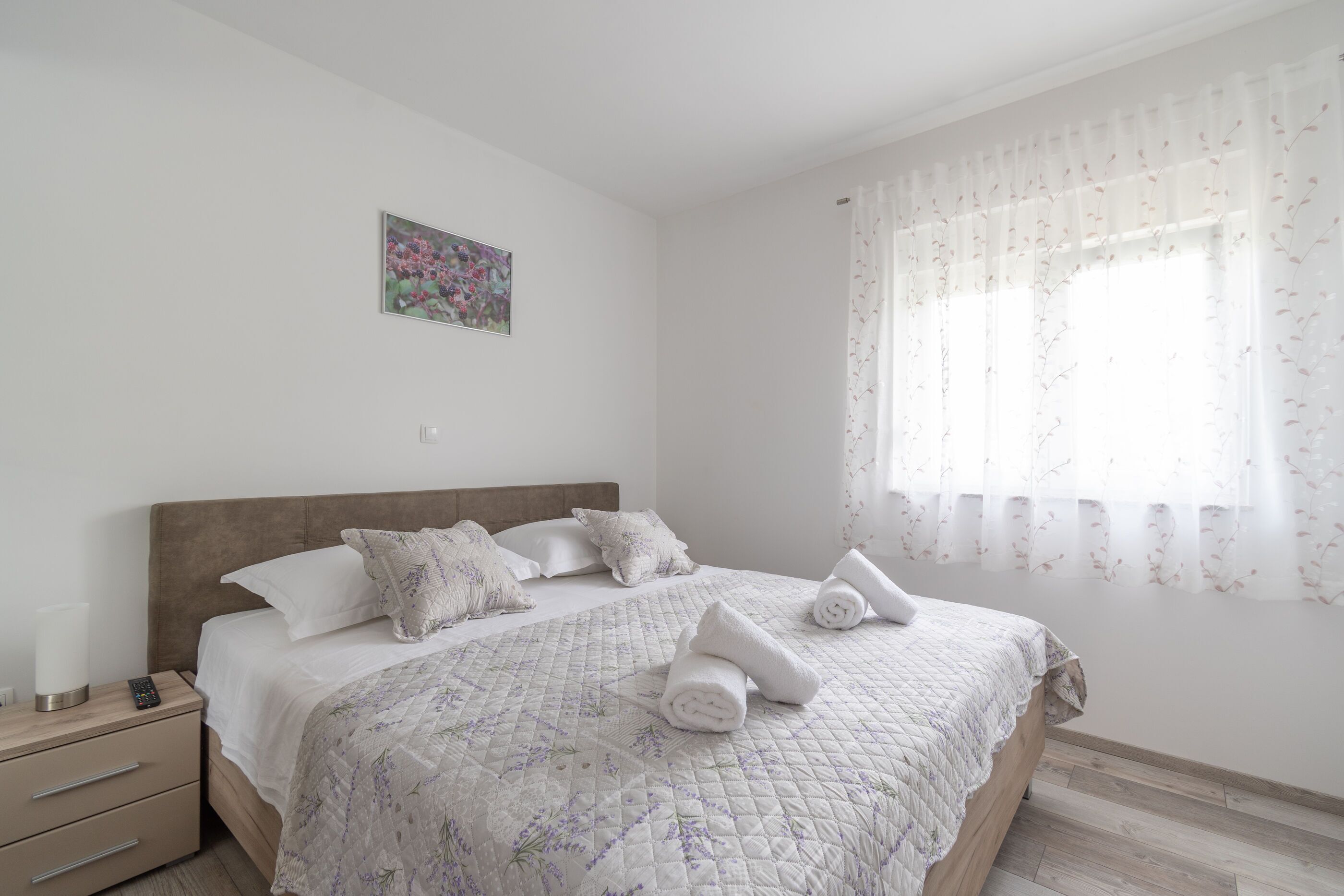 A neatly made bed with a floral quilt, a bedside table with a lamp, and a window with sheer curtains.