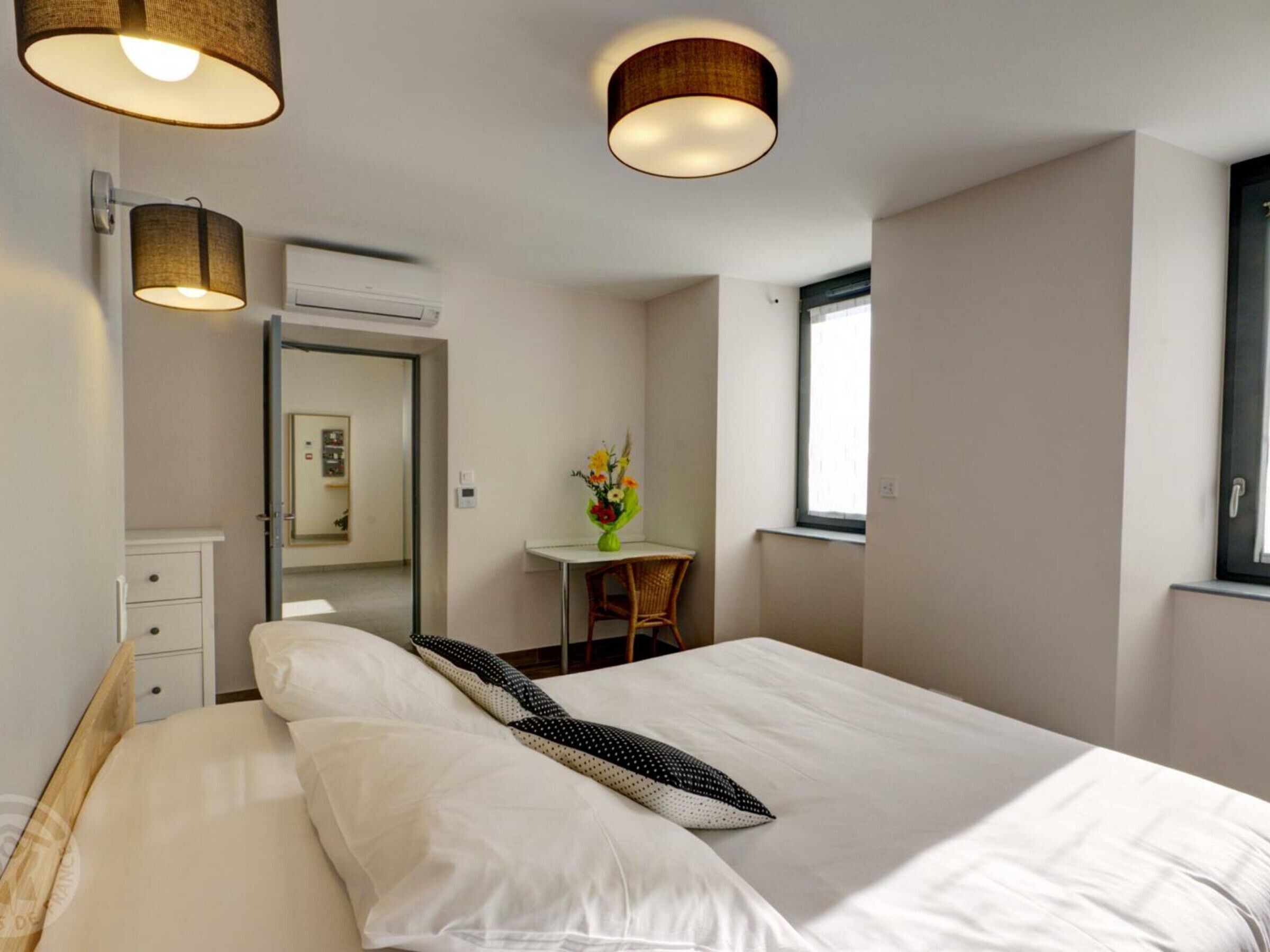 Une chambre d’hôtel moderne dotée d’un grand lit, de lampes de chevet, d’un petit bureau avec une chaise et d’une fenêtre laissant entrer la lumière naturelle.