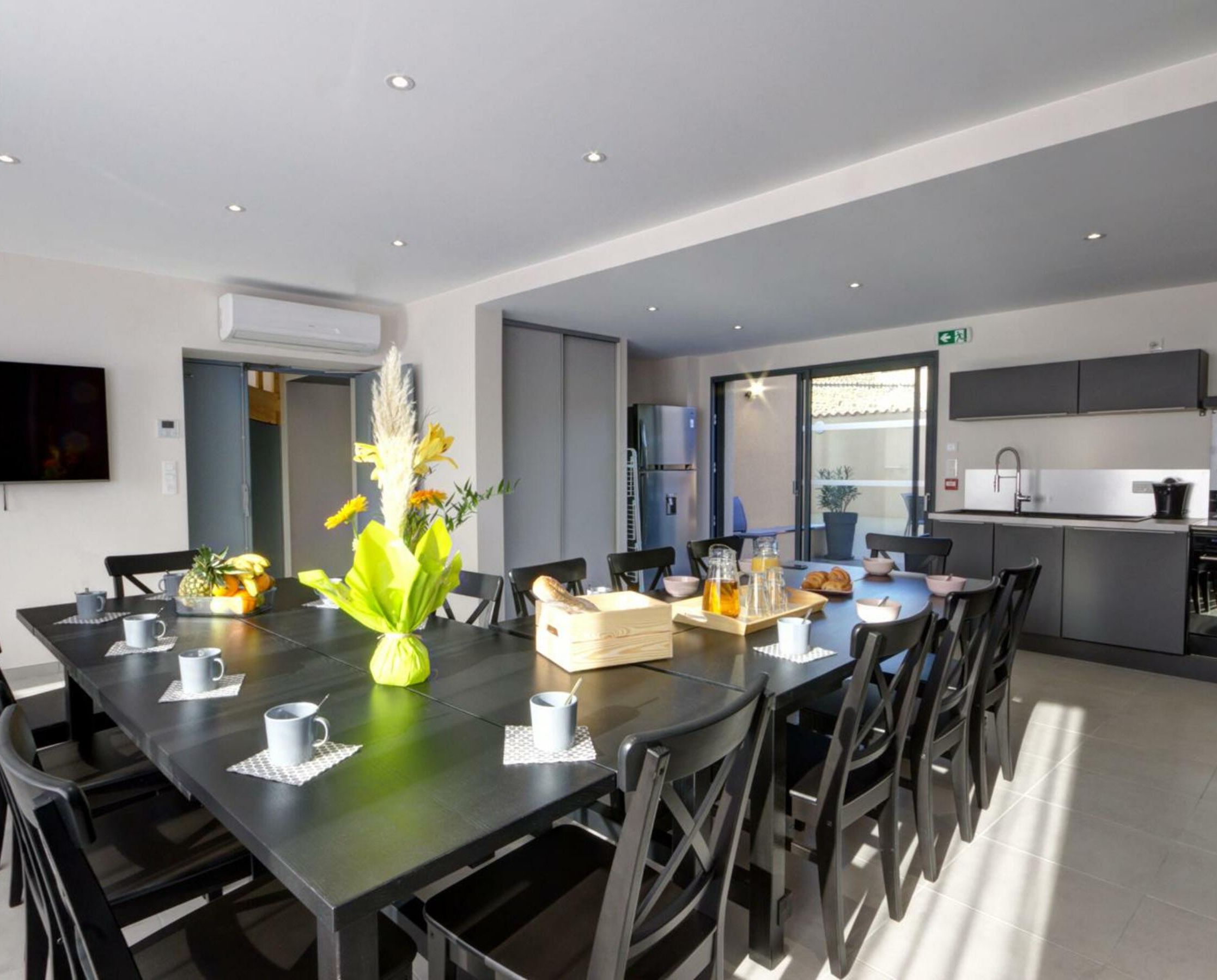A modern dining area with a long table, chairs, and a vase of flowers. There is a kitchen area with cabinets and a sink visible in the background.