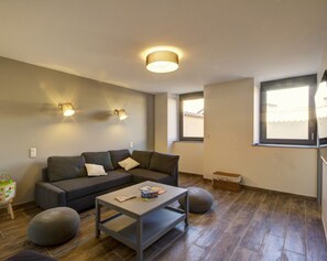 Una sala de estar moderna con un sofá seccional gris, una pequeña mesa de centro de madera y un taburete gris. Hay dos lámparas de pared y una lámpara de techo.