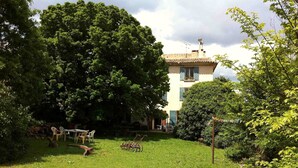 Un jardín exuberante con un árbol grande, una pequeña mesa con sillas y un edificio de techo a la vista.