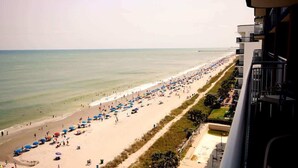 A beach with umbrellas and people, a clear sky, and a building with balconies. - Heaven on Earth Summa (Myrtle Beach)
