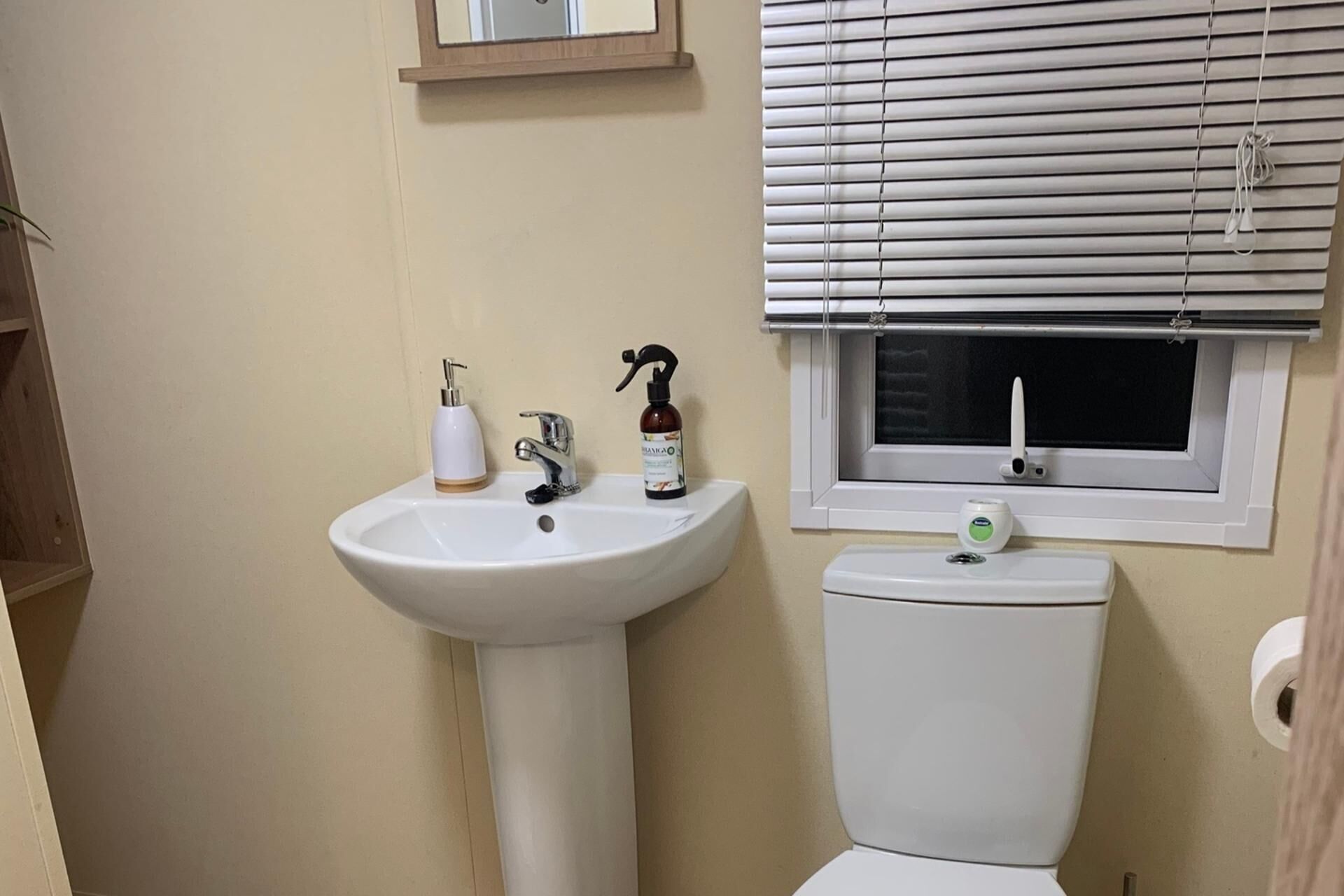 Une salle de bain avec un lavabo blanc, des toilettes et une fenêtre avec des stores.