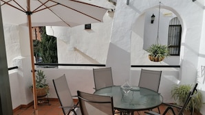 Un tavolo rotondo in vetro con quattro sedie, un portavaso sospeso e una piccola pianta in vaso su un patio piastrellato con pareti e archi bianchi.