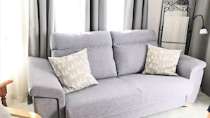 Un divano grigio con due cuscini decorati, una lampada da terra e tende in una stanza con pavimento piastrellato.