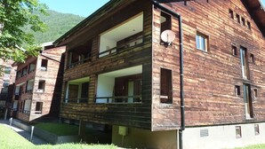 Ein zweistöckiges Holzhaus mit einem Balkon im oberen Stockwerk, großen Fenstern und einer Satellitenschüssel.