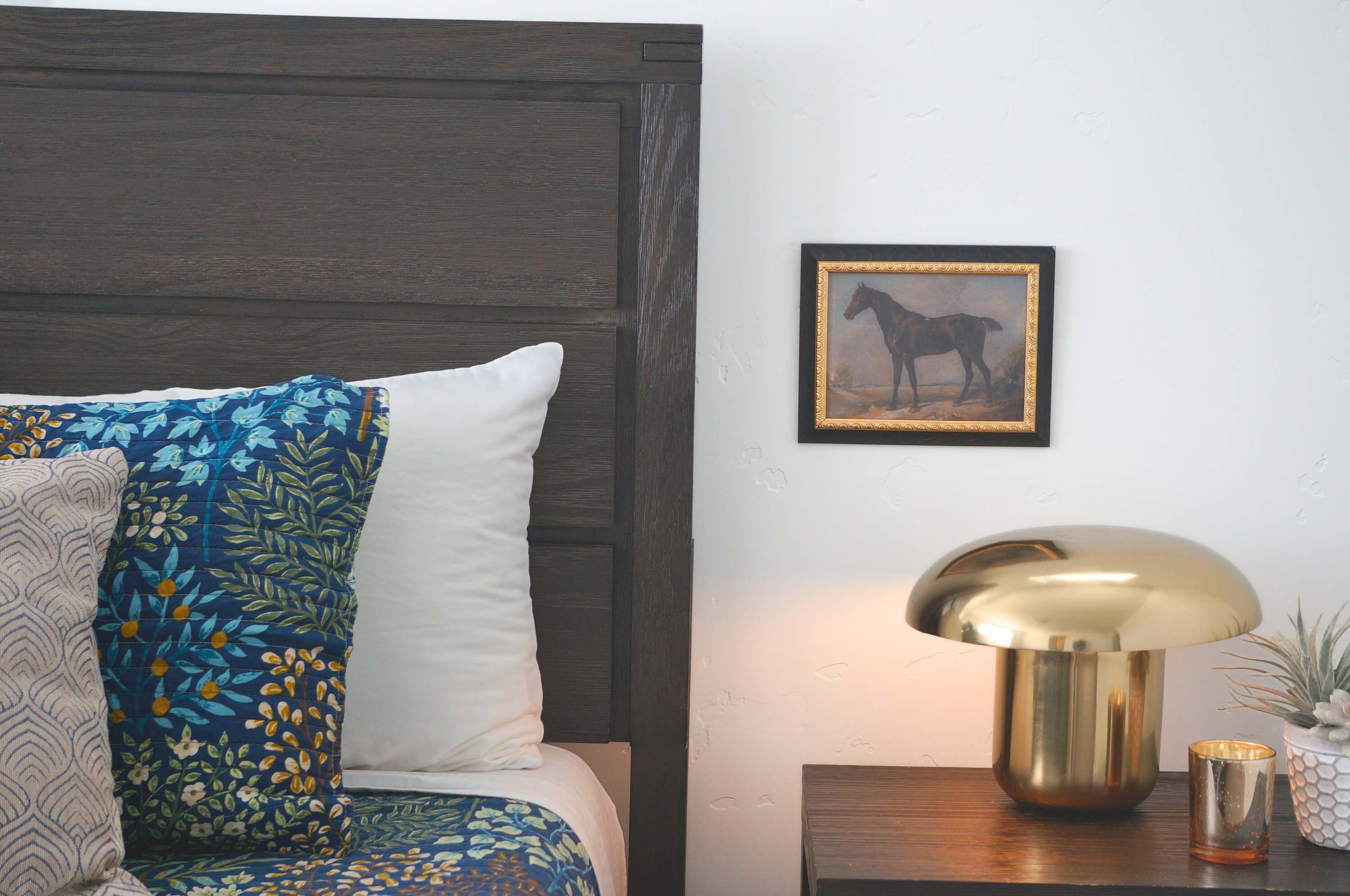 A bedroom with a bed, pillows, a framed picture, a lamp, and a small table with a plant.