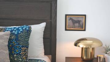 A bedroom with a bed, pillows, a framed picture, a lamp, and a small table with a plant.