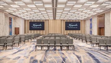 Un salón de conferencias amplio con filas de sillas, una alfombra estampada y dos pantallas grandes mostrando el nombre del hotel.