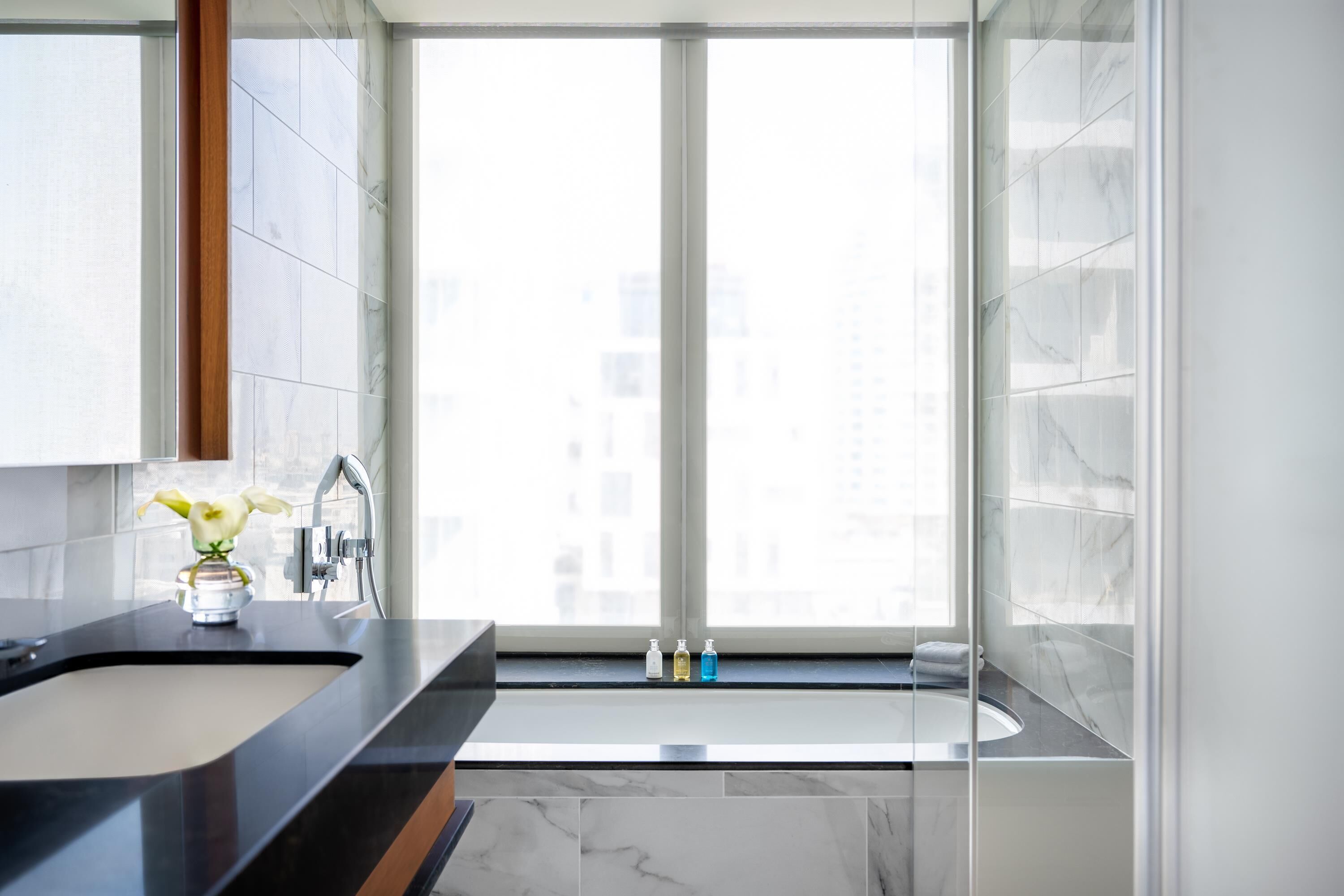 A modern bathroom with a large window, a black countertop, a white sink, and a vase with flowers.