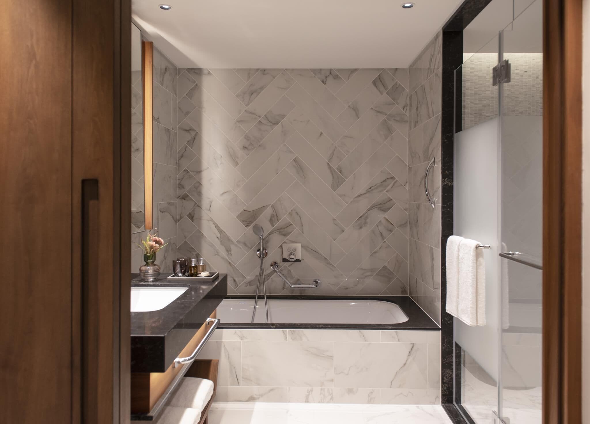 A modern bathroom with a marble herringbone wall pattern, a black bathtub, a glass shower enclosure, and a wooden vanity with a mirror.