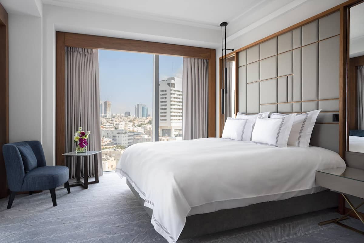 Une chambre d’hôtel moderne avec un grand lit, une petite table ornée d’un vase de fleurs et une vue sur le paysage urbain grâce à de grandes fenêtres.