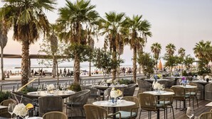 An outdoor dining area with round tables and wicker chairs, overlooking a beach with palm trees and a view of the ocean. - The David Kempinski Tel Aviv (Tel Aviv)