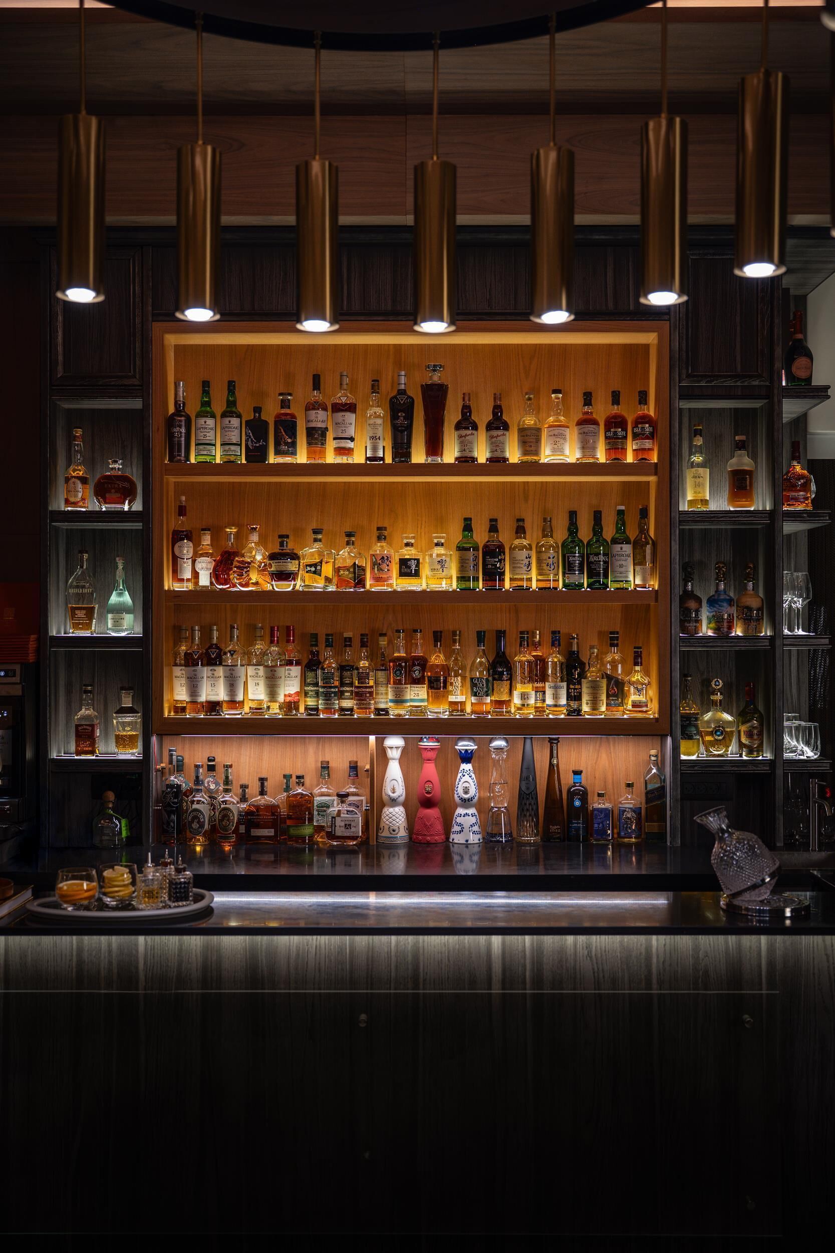 Zona de bar con varias estanterías llenas de botellas de licor, lámparas colgantes y una barra.