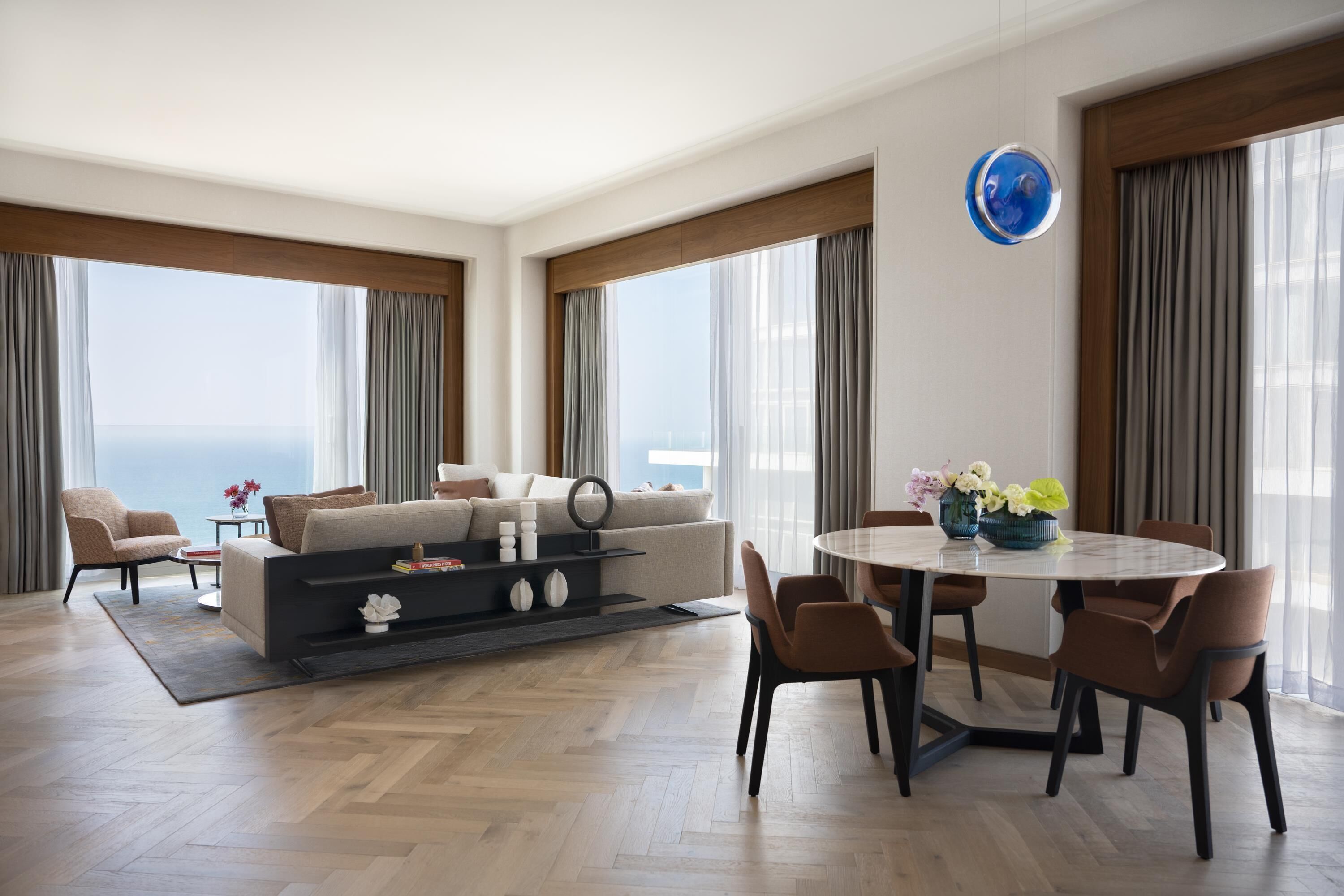 Un salon moderne avec un canapé, une table basse et un coin repas, de grandes fenêtres avec des rideaux et une vue sur l’océan.