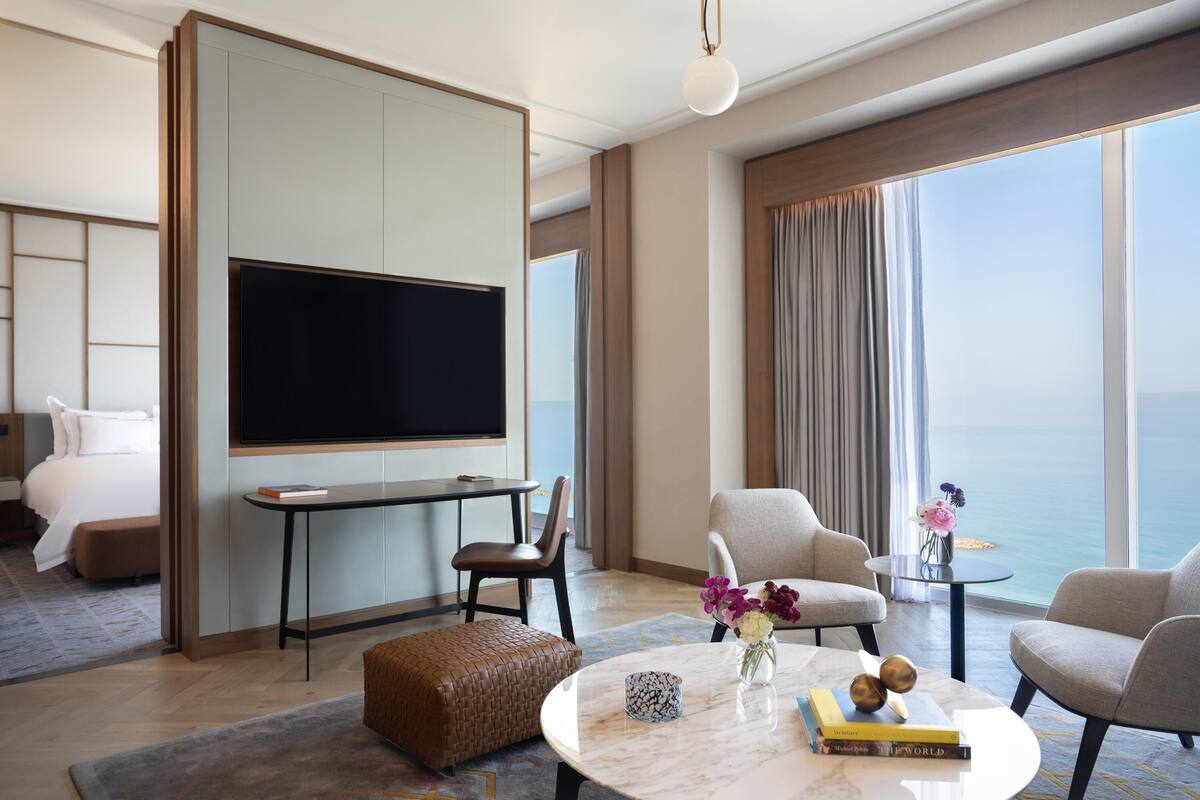 Une chambre d’hôtel moderne dotée d’une grande télévision à écran plat, de fauteuils confortables, d’une petite table ornée d’un vase de fleurs et offrant une vue sur l’océan.