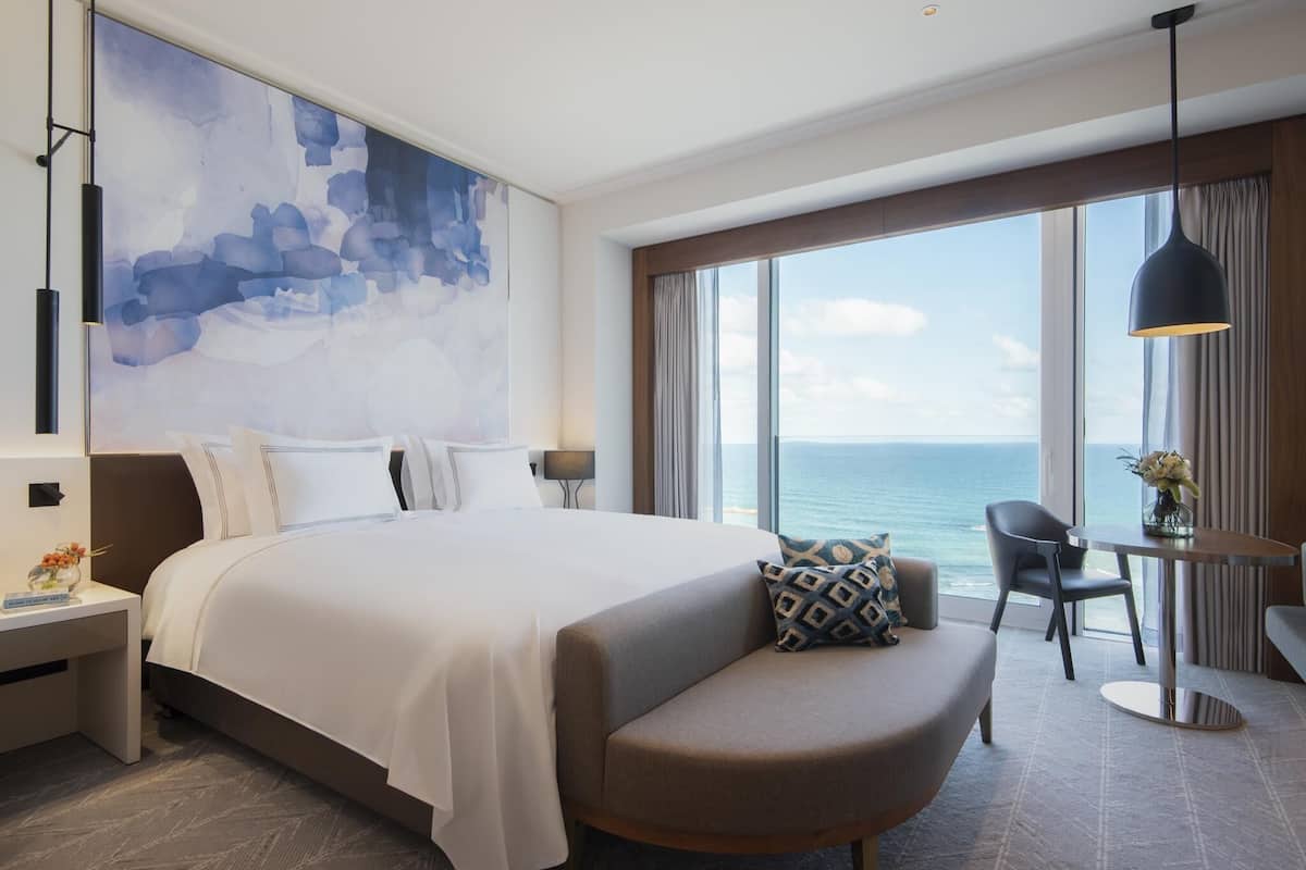 Une chambre d’hôtel moderne dotée d’un grand lit, d’un coin salon, d’une table à manger et d’une grande fenêtre offrant une vue sur l’océan.