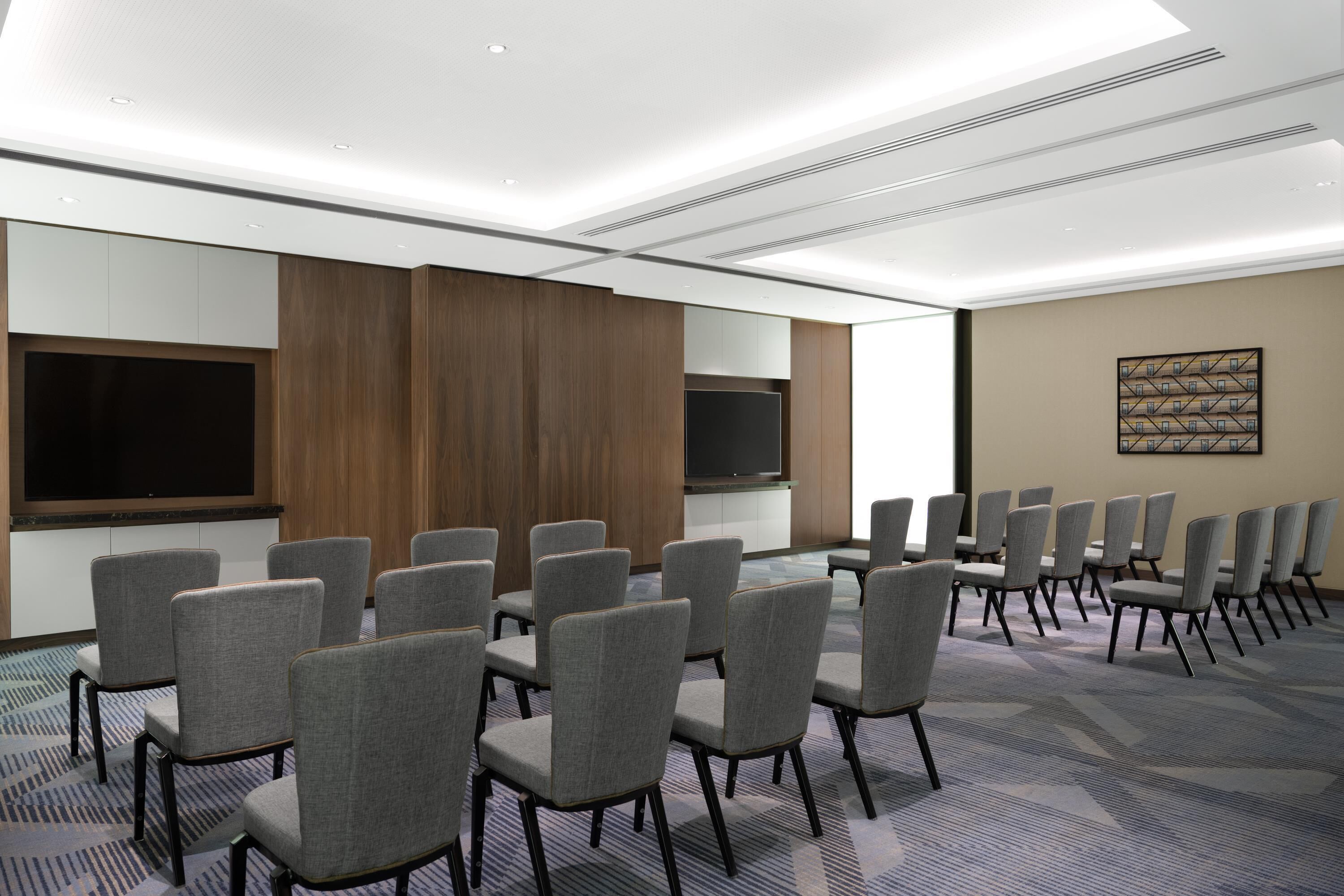 A conference room with rows of grey chairs, a large flat-screen TV, wooden paneling, and a patterned carpet.