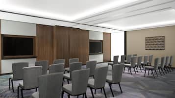 A conference room with rows of grey chairs, a large flat-screen TV, wooden paneling, and a patterned carpet.