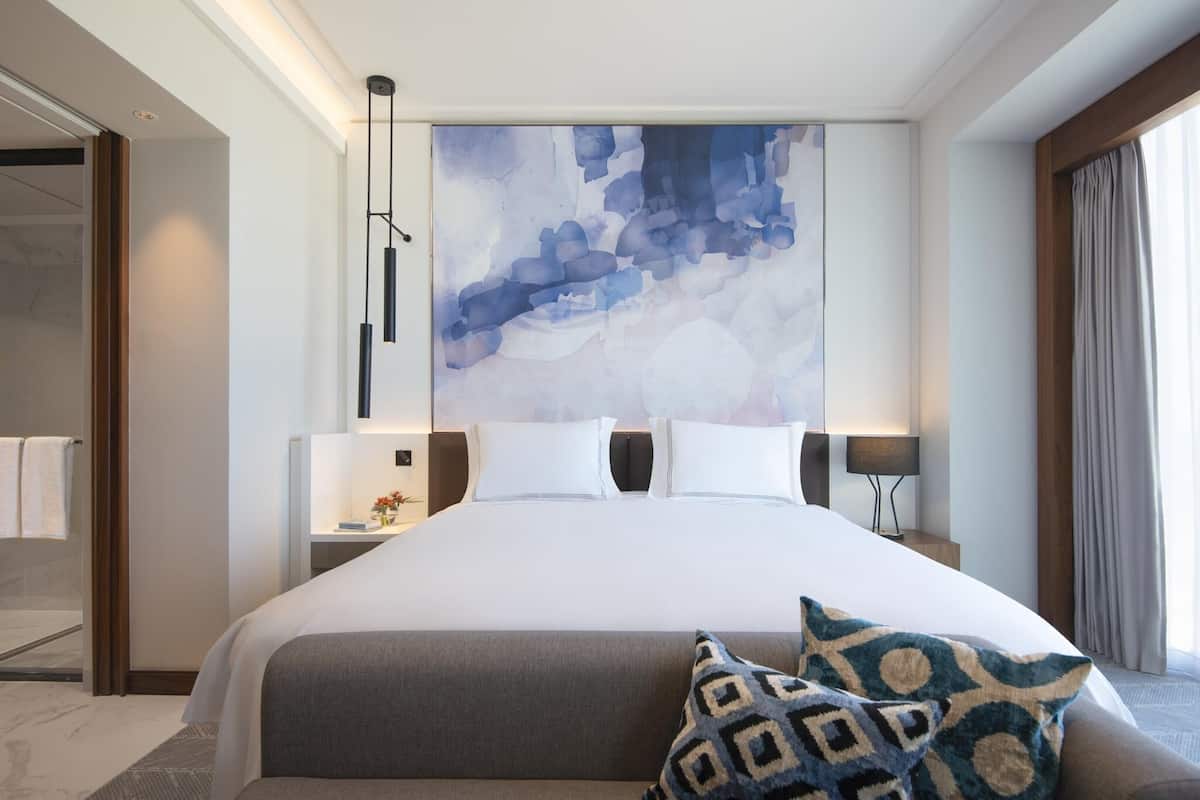 Une chambre d’hôtel moderne avec un grand lit, un tableau abstrait accroché au mur et une vue sur l’extérieur par une fenêtre.