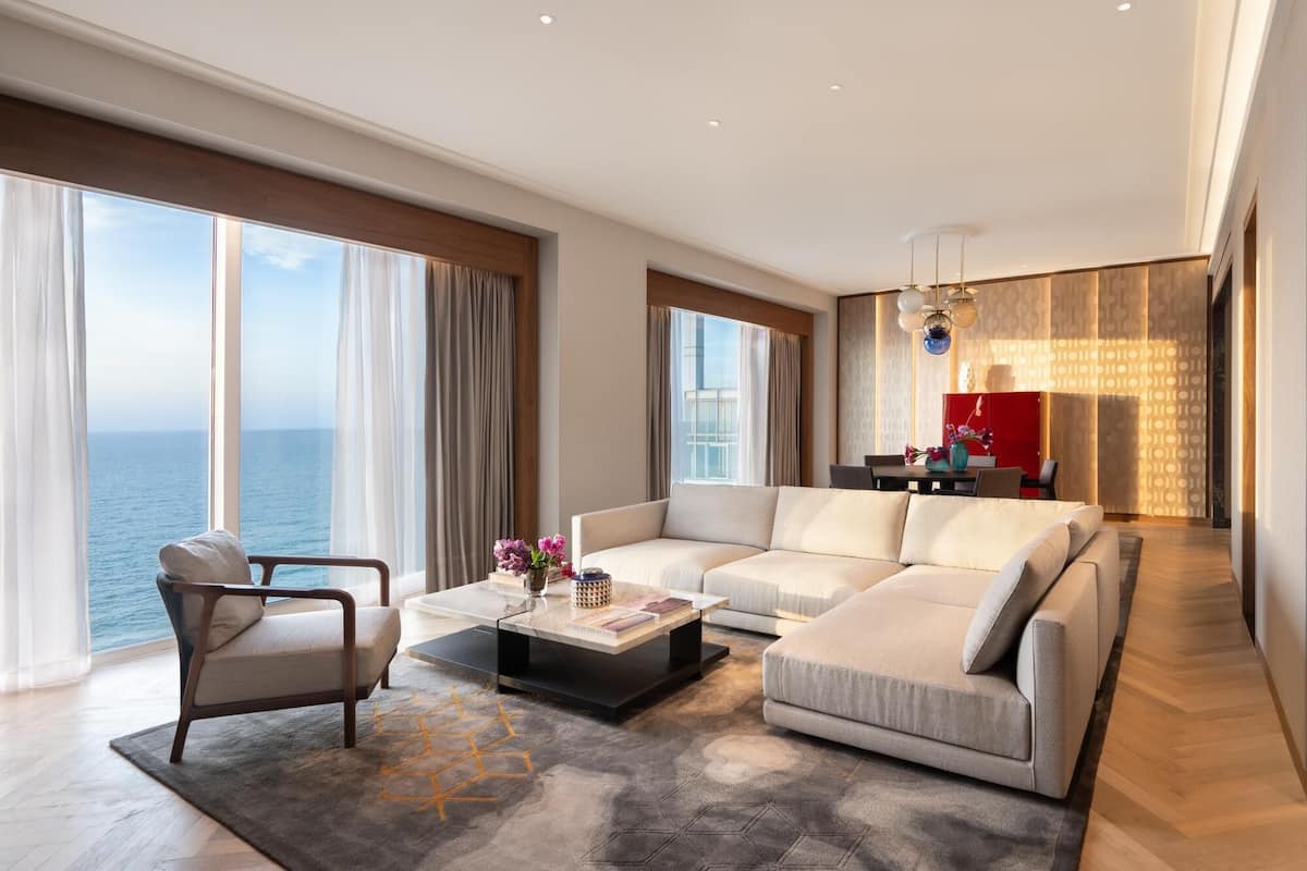Un salon moderne avec un grand canapé, une table basse et un fauteuil. De grandes fenêtres offrent une vue sur l’océan.