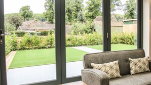 Une pièce avec un canapé gris, deux coussins à motifs et une petite table en bois. Une grande fenêtre offre une vue sur un jardin bien entretenu et un quartier résidentiel.