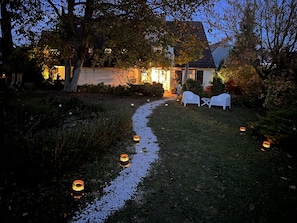 Un sendero iluminado con velas que conduce a una casa de paredes blancas y techo oscuro. Hay dos sillas de plástico blancas fuera de la casa.