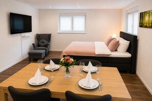 Habitación de hotel con cama, televisión, mesa de comedor para cuatro personas y ventana con persianas.