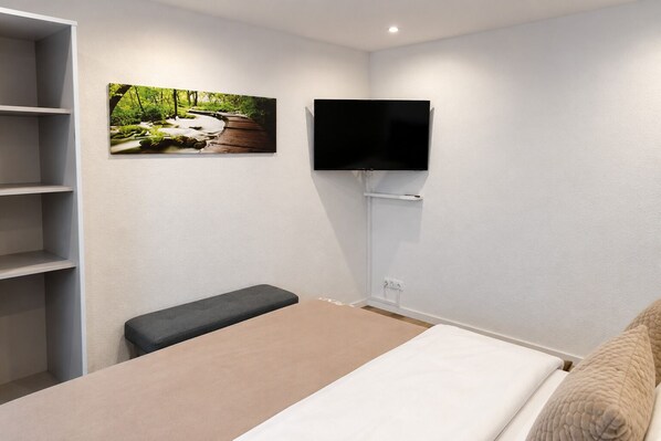 Un dormitorio con cama, una banca, televisión montada y repisa en la pared.