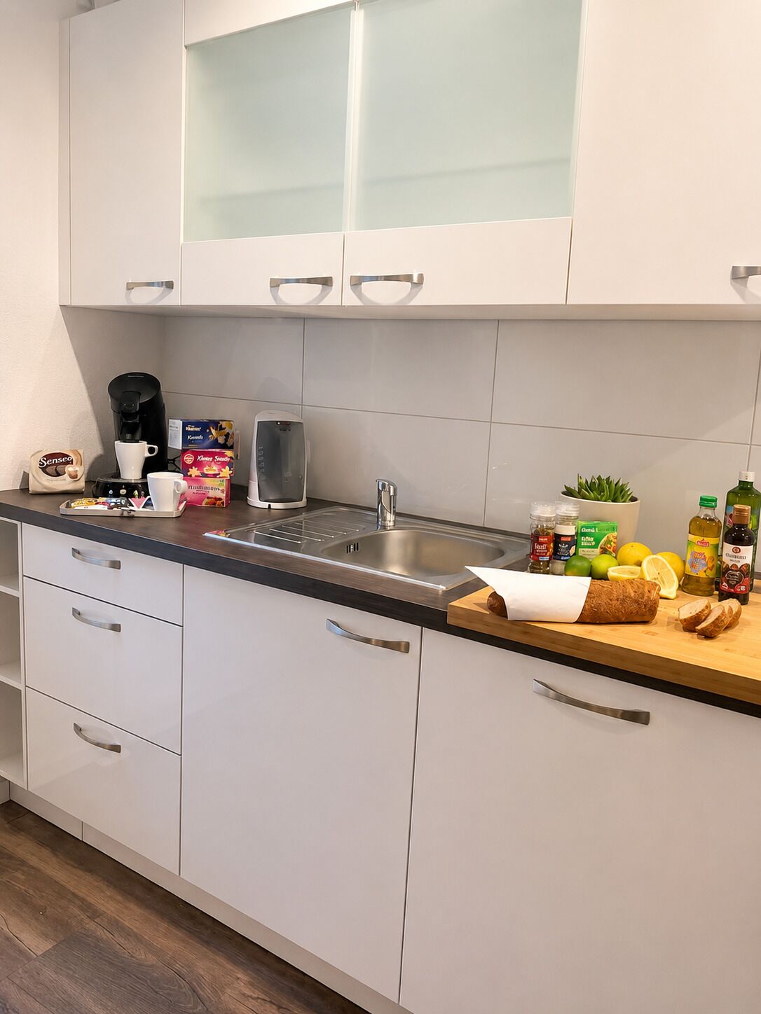 A modern kitchen with white cabinets, a stainless steel sink, and various food items on the counter.