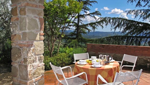 An outdoor dining area with a stone pillar, white metal chairs, and a table set with dishes and a pitcher. - Casa Citille (Greve in Chianti)