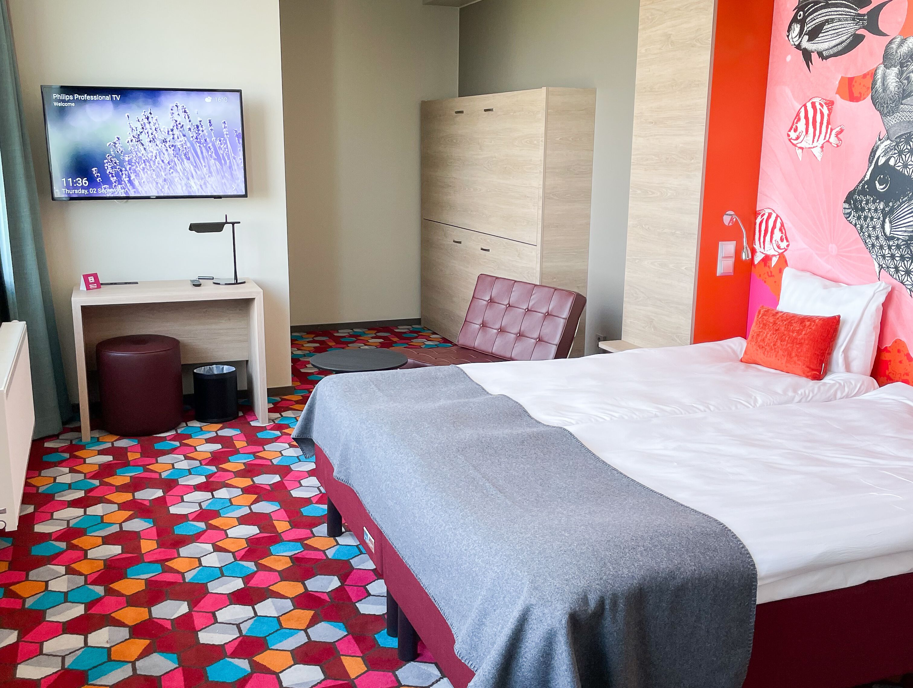 Habitación de hotel con cama, escritorio, silla, armario y televisor mostrando una pantalla de bienvenida.
