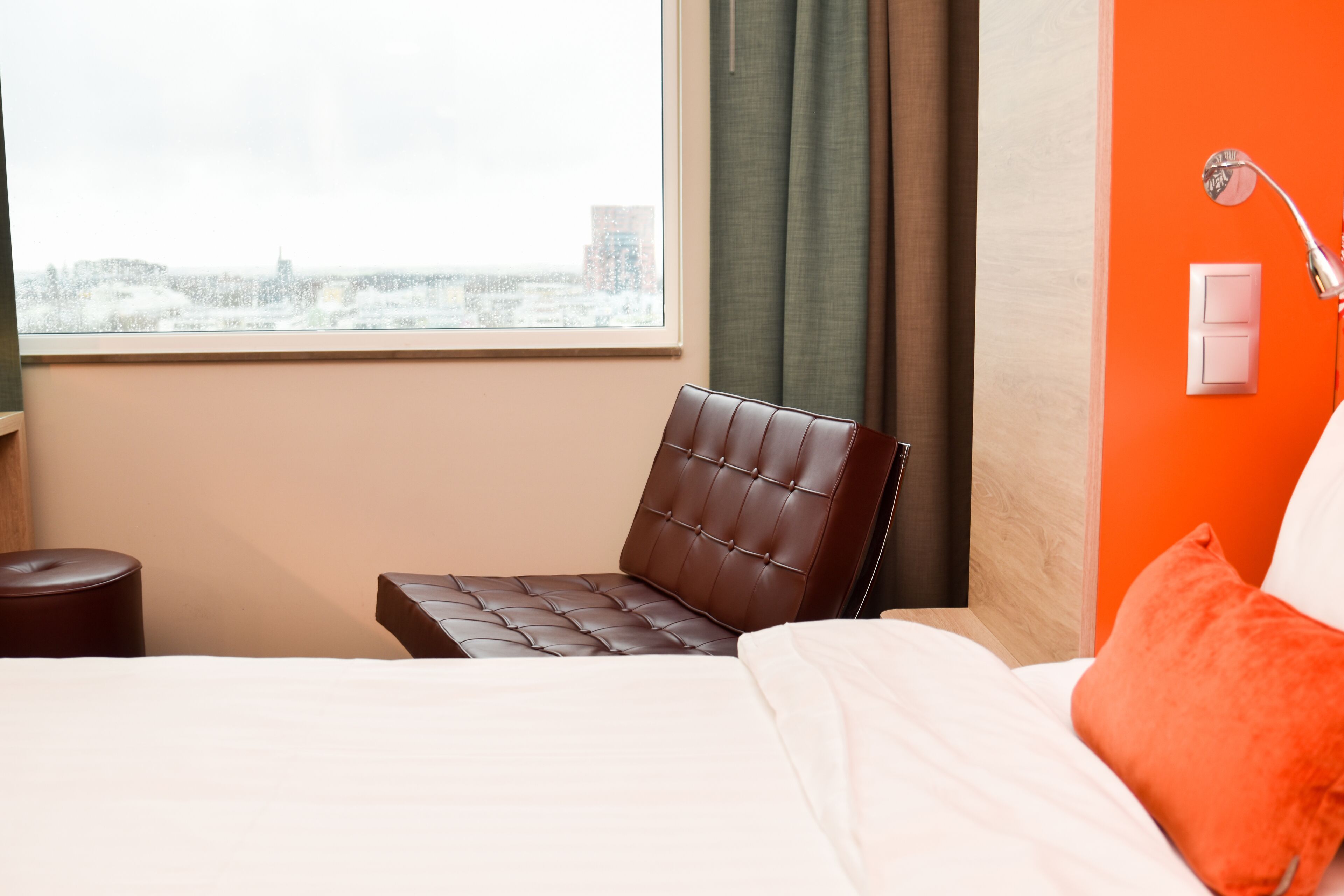 Een hotelkamer met een bed, een bruine leren fauteuil, een raam met uitzicht over de stad en een oranje accentmuur.