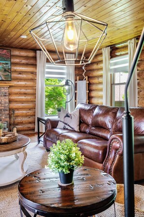 A cozy log cabin interior with a leather sofa, a round wooden table with a plant, and a hanging light fixture.