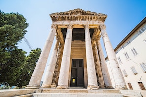 A classical building with columns and a central door, surrounded by trees and adjacent buildings.
