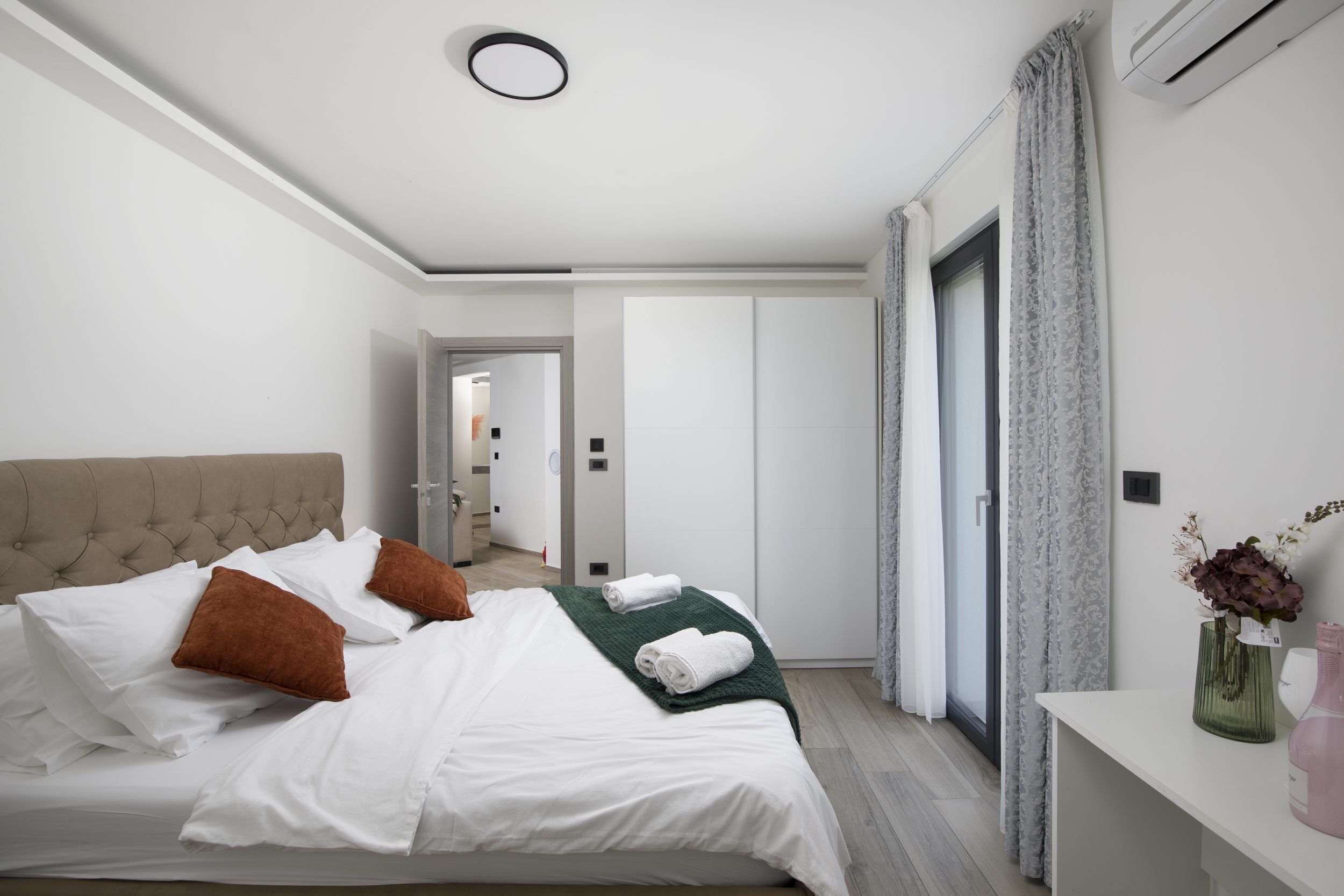 Ein Hotelzimmer mit Bett, Nachttisch mit Vase, Tür und Flur.