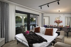 A modern hotel room with a large bed, a dining area, and a view of the outdoors.
