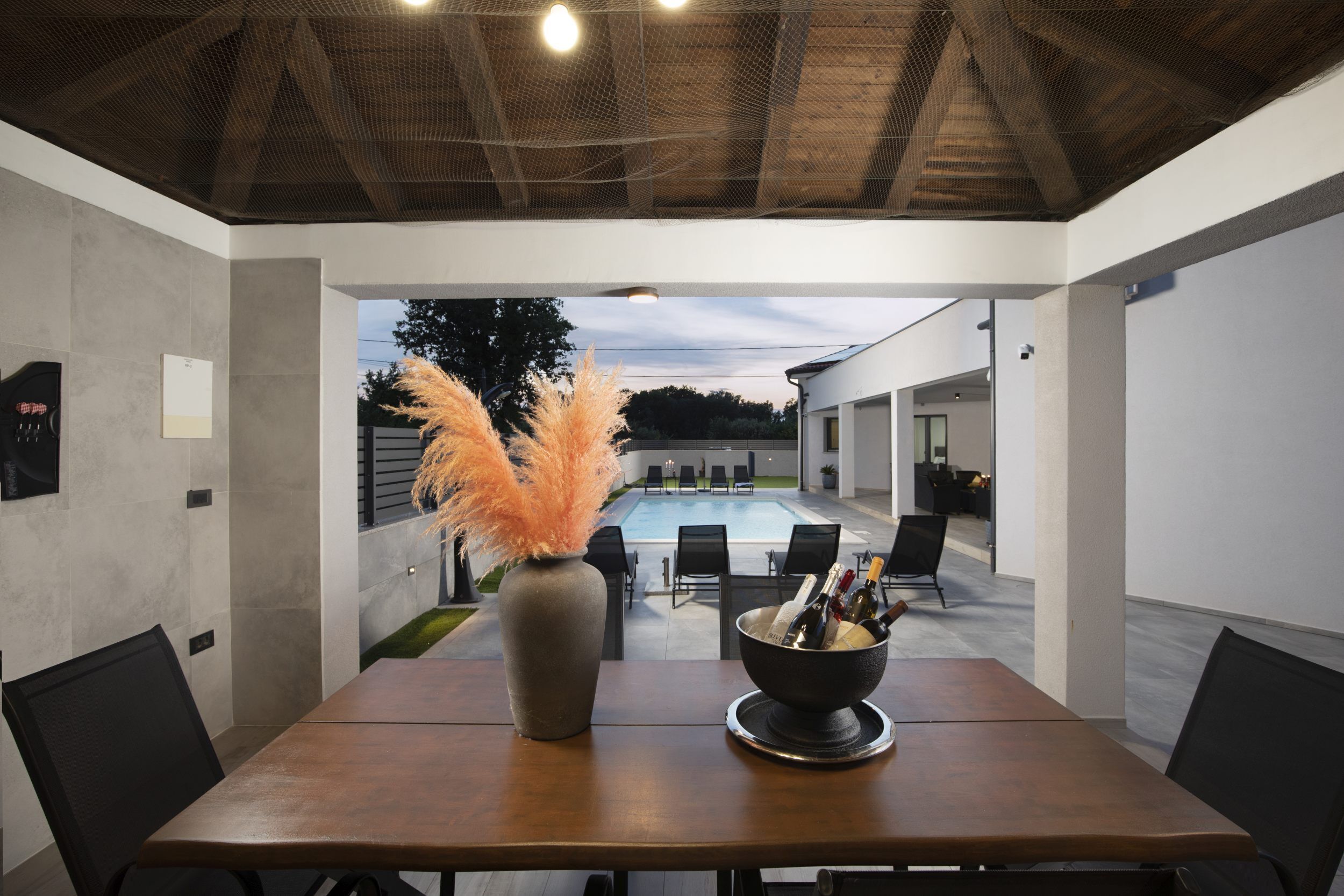 Un moderno comedor con una mesa, sillas, un jarrón con plumas de miscanthus y vistas a una piscina y mobiliario exterior.