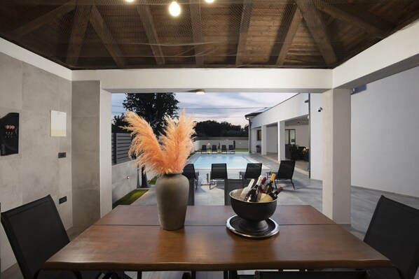 A modern dining area with a table, chairs, a vase with pampas grass, and a view of a swimming pool and outdoor seating. - Villa Speranza with pool (Pula)