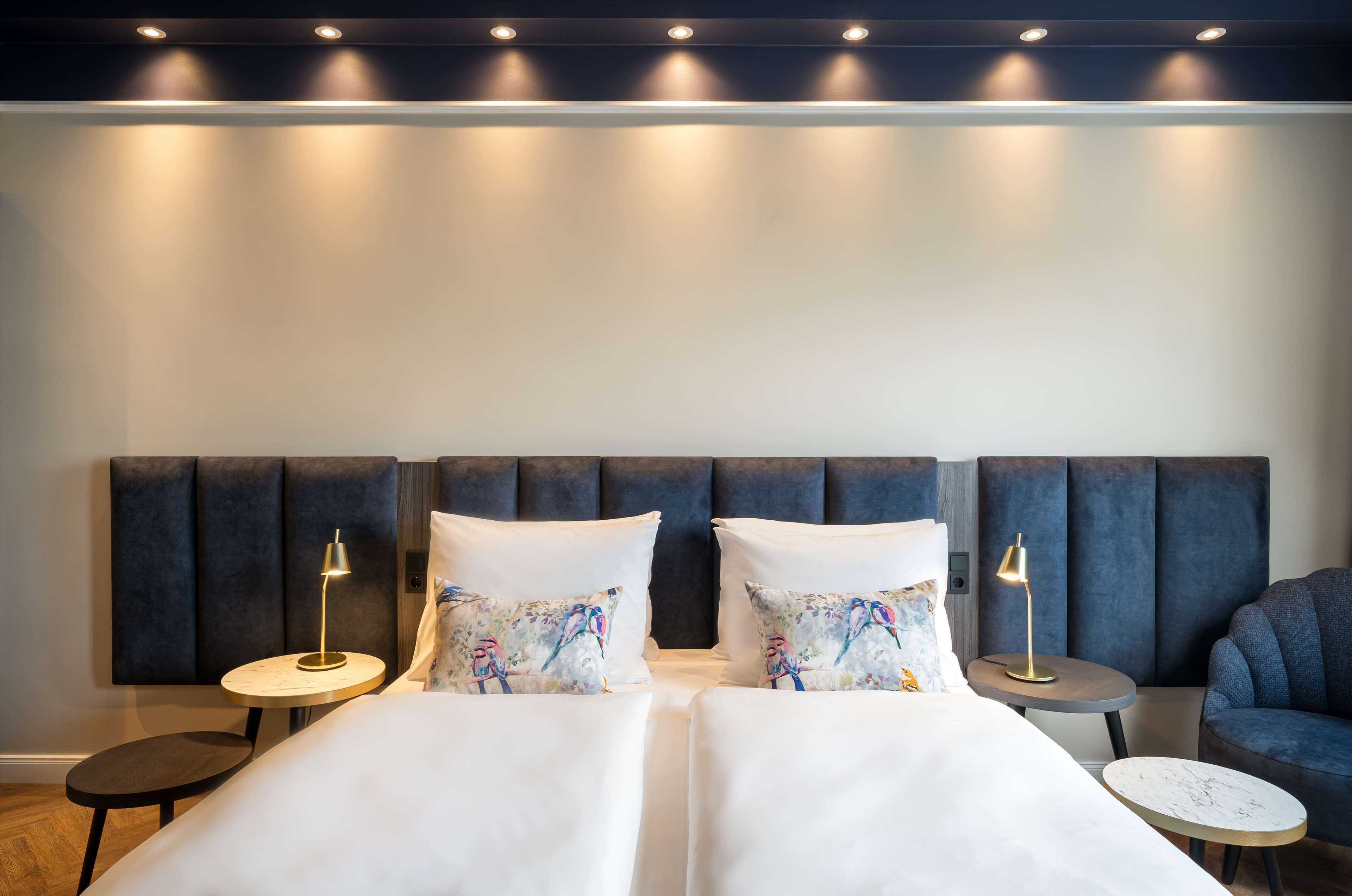 Habitación de hotel con una cama grande, dos mesitas de noche con lámparas, una mesita redonda pequeña y un sillón azul.