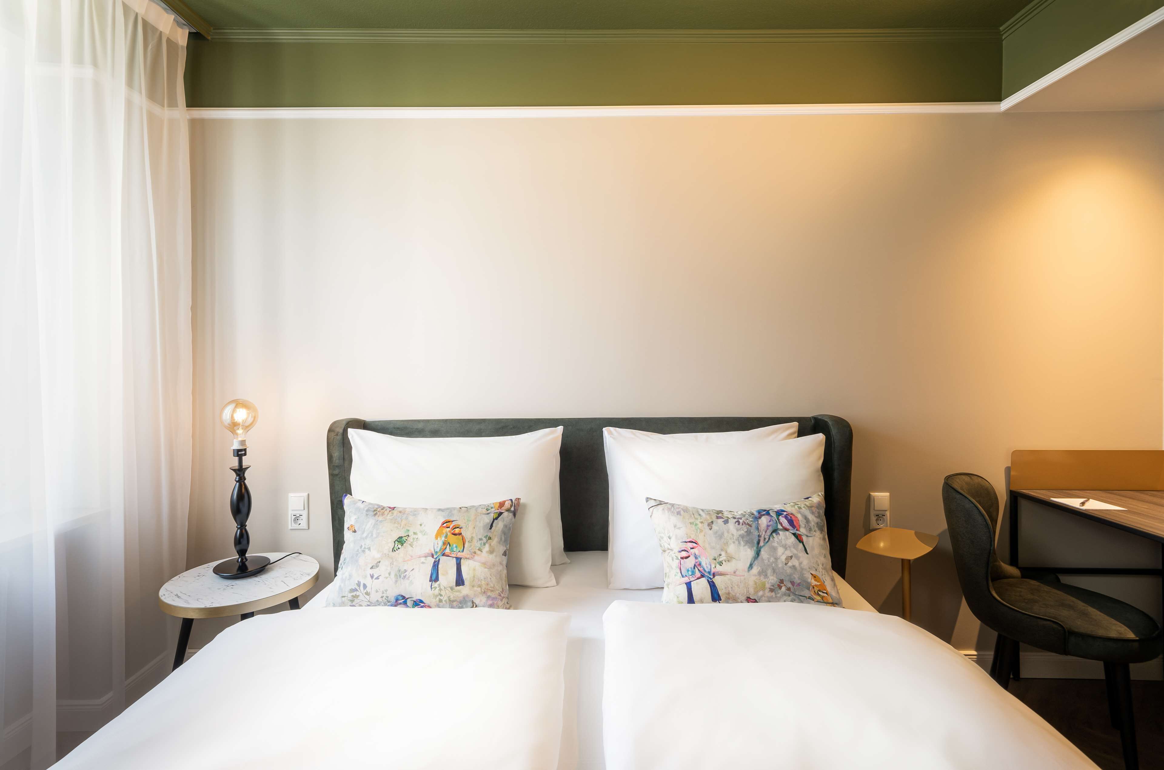 A hotel room with a double bed, two decorative pillows, a bedside table with a lamp, a chair, and a desk.