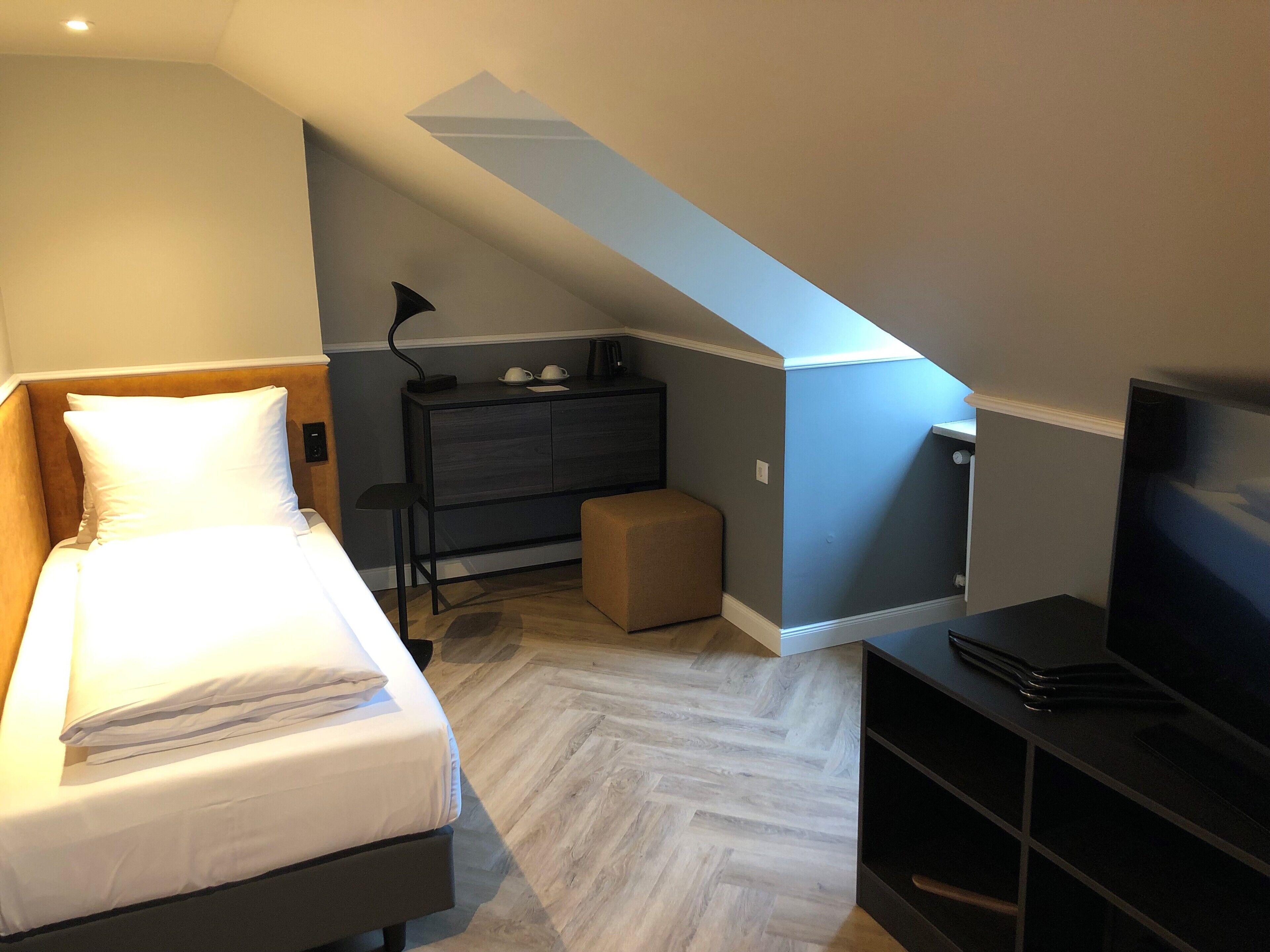 A bedroom with a bed, a desk, a chair, a TV, and a small ottoman.