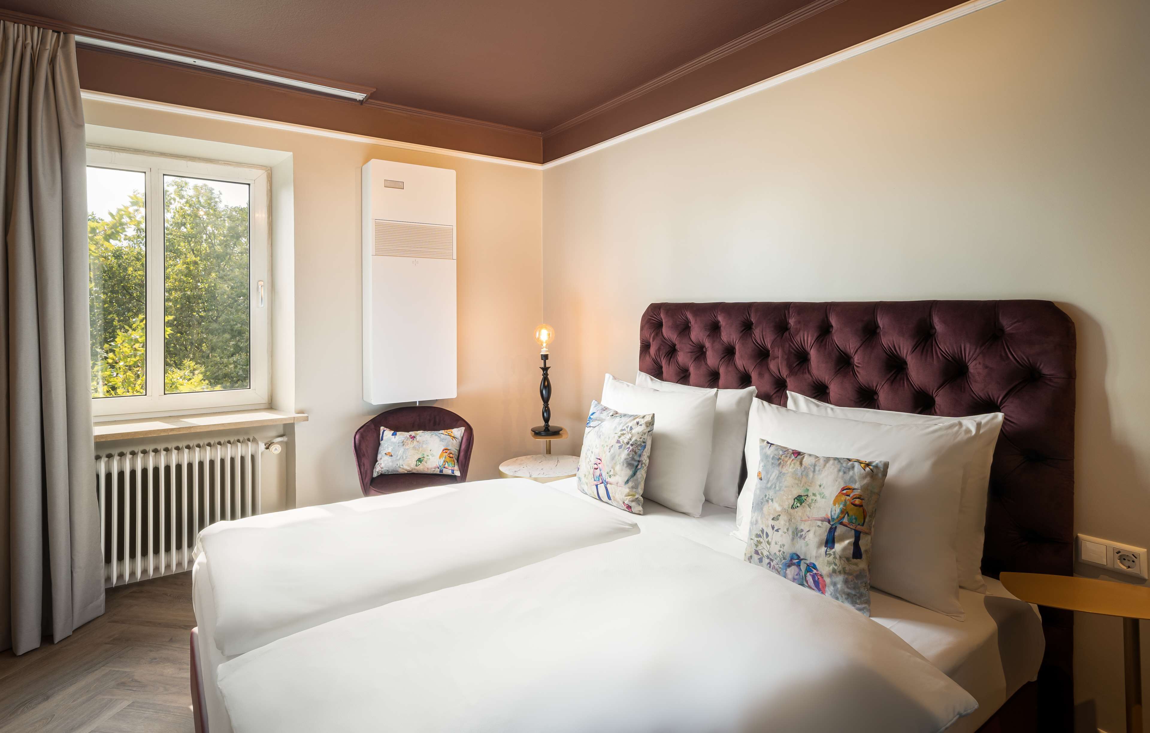 Habitación de hotel con una cama grande, una ventana con cortinas, un radiador, una mesita de noche con lámpara y una silla con cojín.