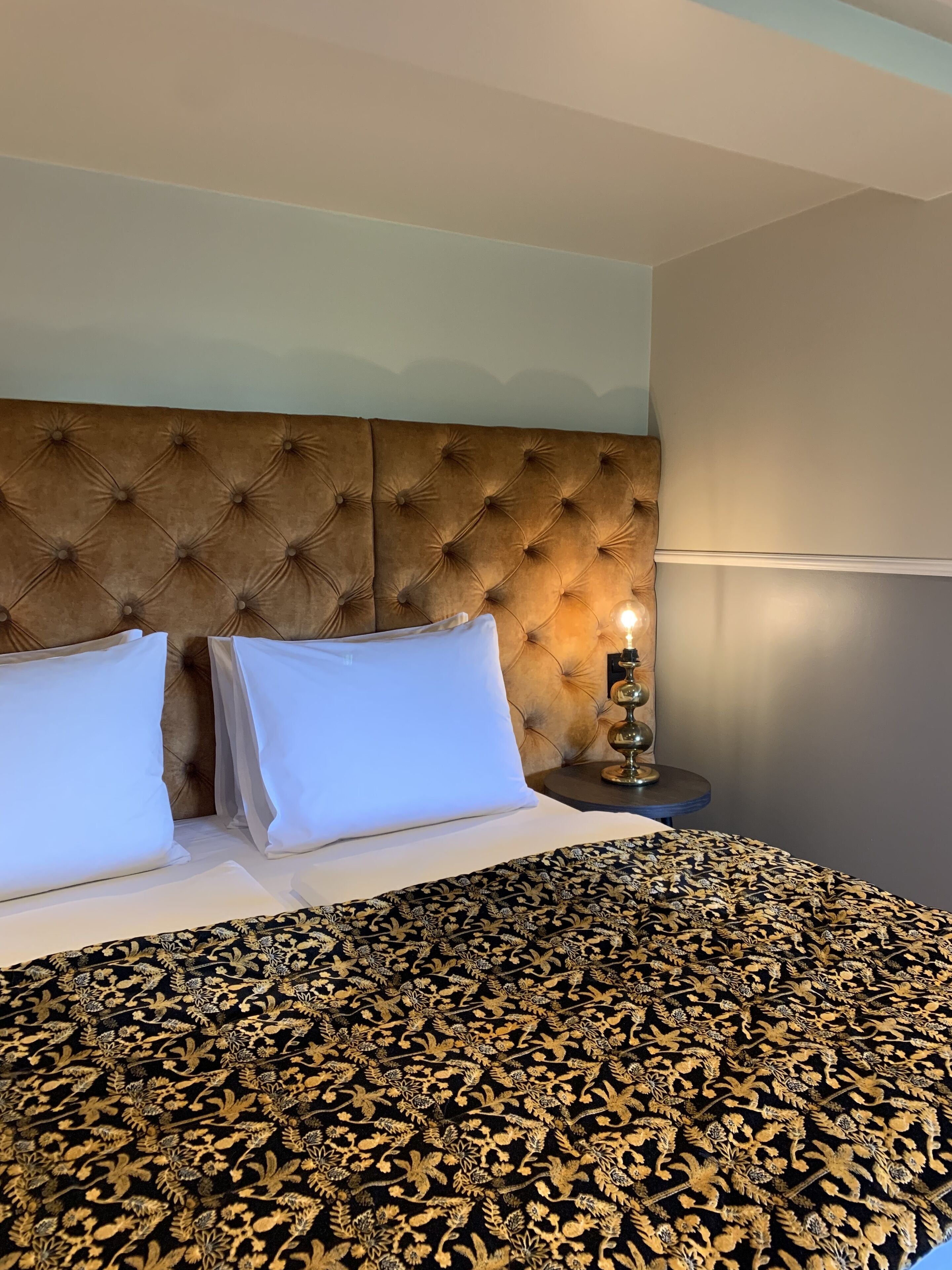 Ett hotellrum med en stoppad sänggavel, vita kuddar, ett mönstrat överkast och en sänglampa.