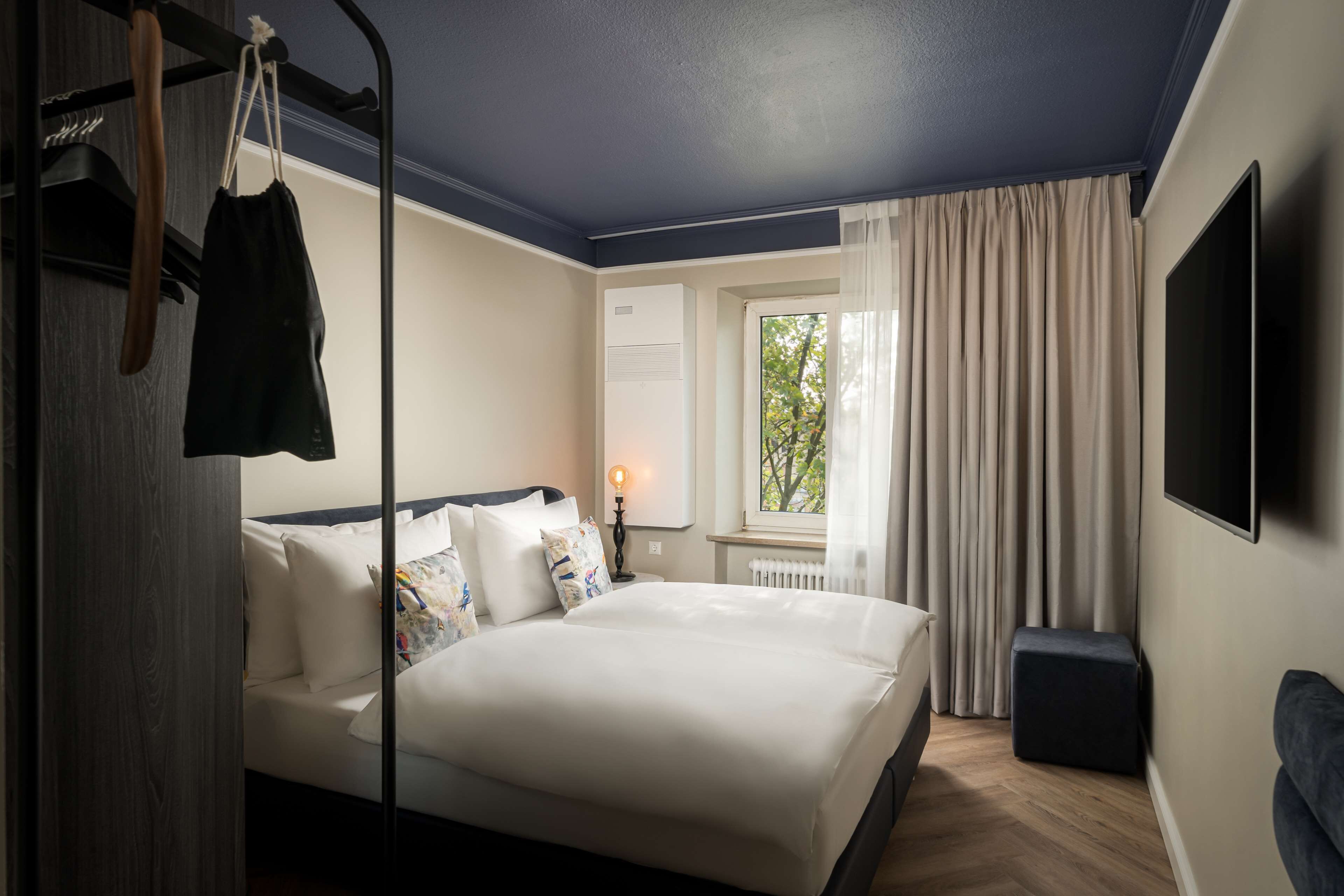 Una habitación de hotel con cama, televisión, ventana con cortinas y radiador.