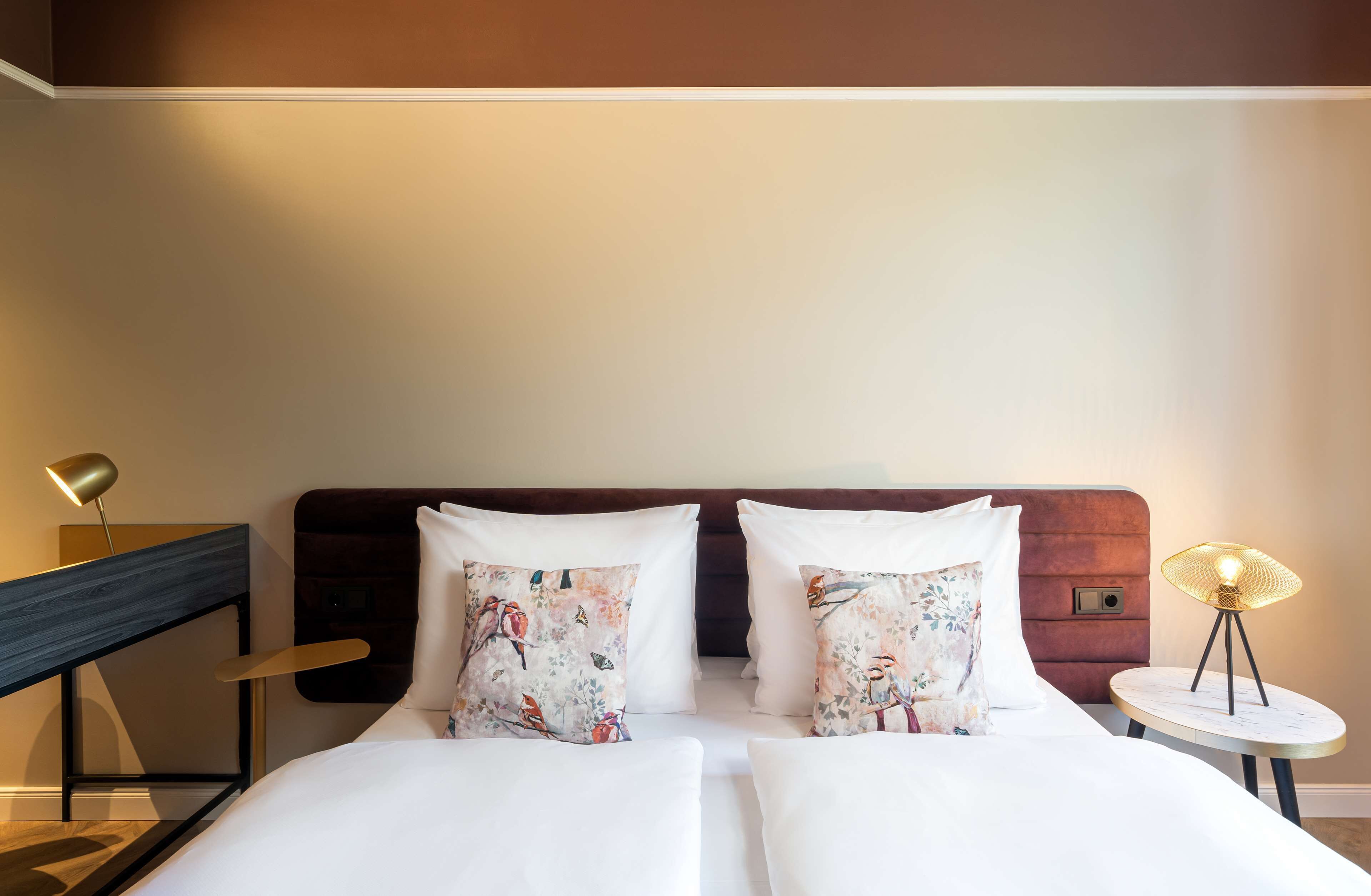 Une chambre d’hôtel avec un lit, deux oreillers à motifs floraux, une table de chevet avec une lampe et une petite table ronde également dotée d’une lampe.