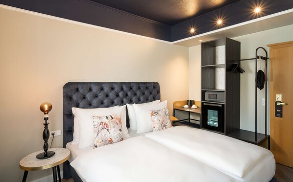 Ein Hotelzimmer mit einem großen Bett, einem Nachttisch mit Lampe, einem Schreibtisch mit Computer und Kaffeemaschine, einem Kleiderschrank und einem Duschbereich.
